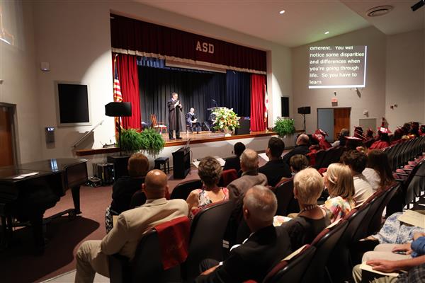 The commencement speaker for Alabama School for the Deaf signs while on stage at graduation. In front of him, the graduates and other audience members look on attentively. 